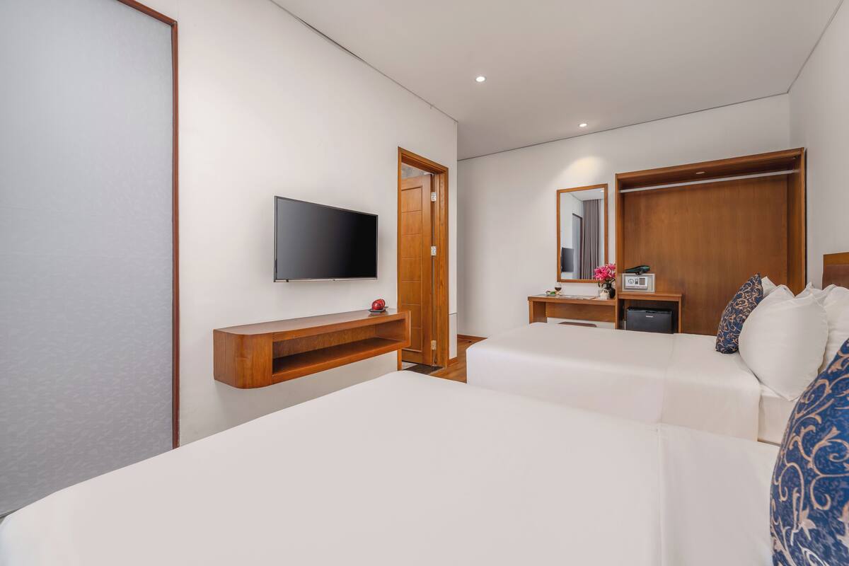 A hotel room with a bed, a TV mounted on the wall, a wooden wardrobe, a bedside table with a lamp, and a mirror.