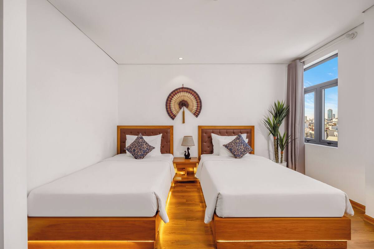 A hotel room with two beds, a wooden floor, a nightstand with a lamp, a wall fan, and a window with a city view.