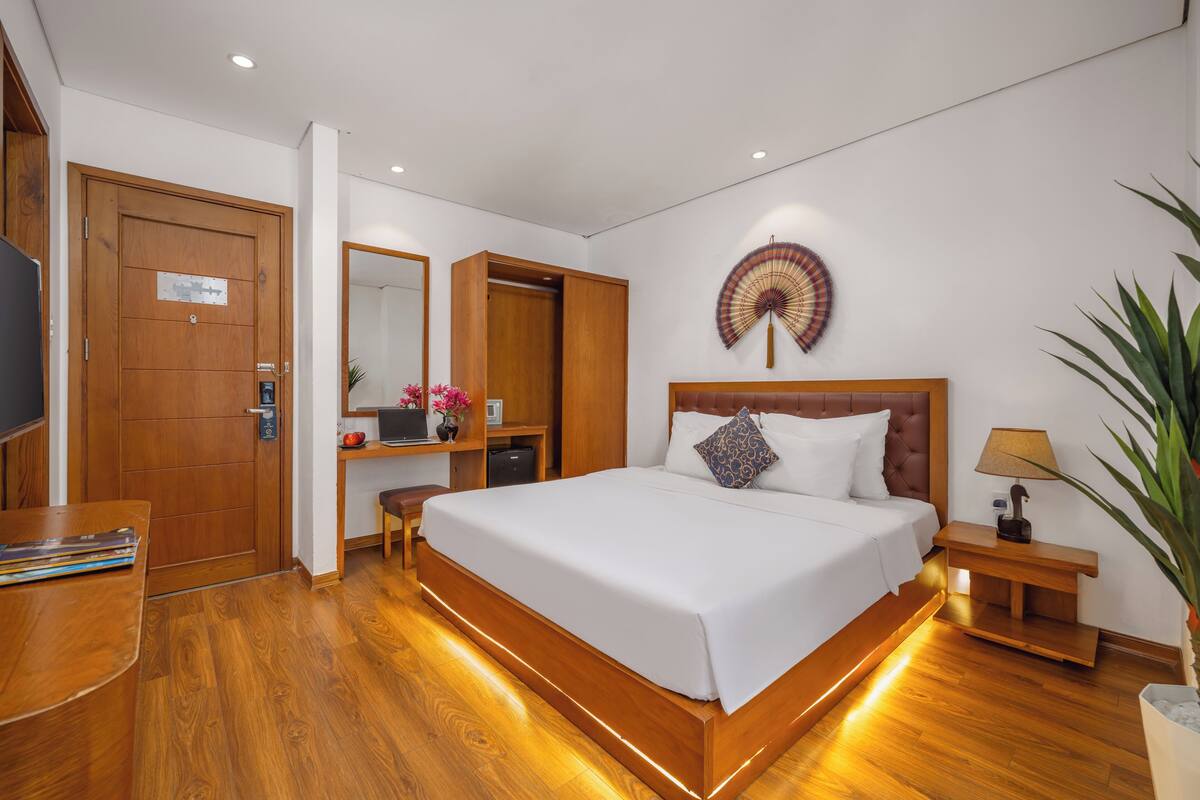 A modern hotel room with a large bed, wooden flooring, a desk with a computer, a TV, and a bedside table with a lamp.