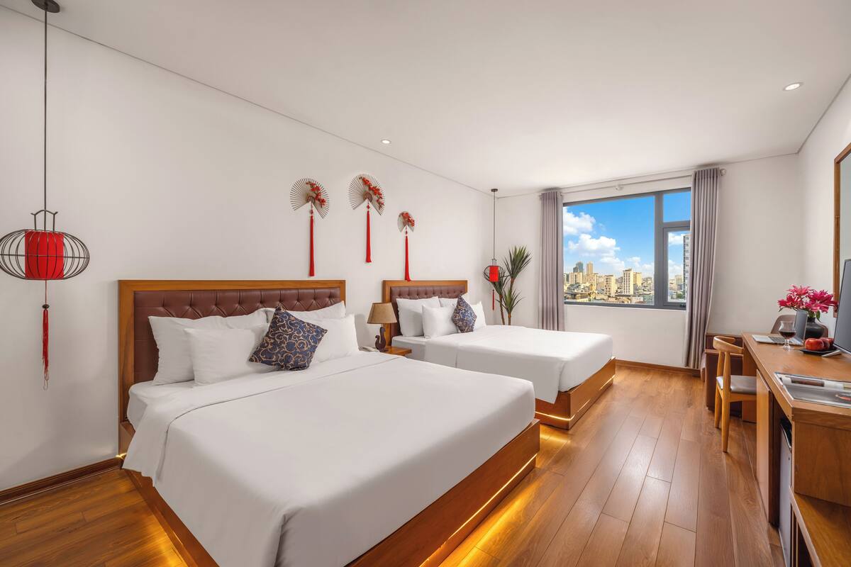 A hotel room with two beds, a desk, and a large window offering a city view.