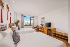 A hotel room with two beds, a desk with a TV, a view of the city, and a large window.