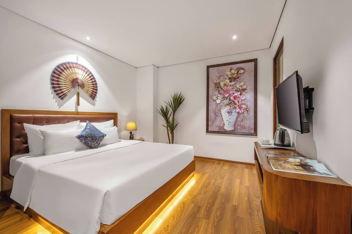 A hotel room with a large bed, a wooden headboard, a bedside table with a lamp, a TV, and a framed floral artwork on the wall.