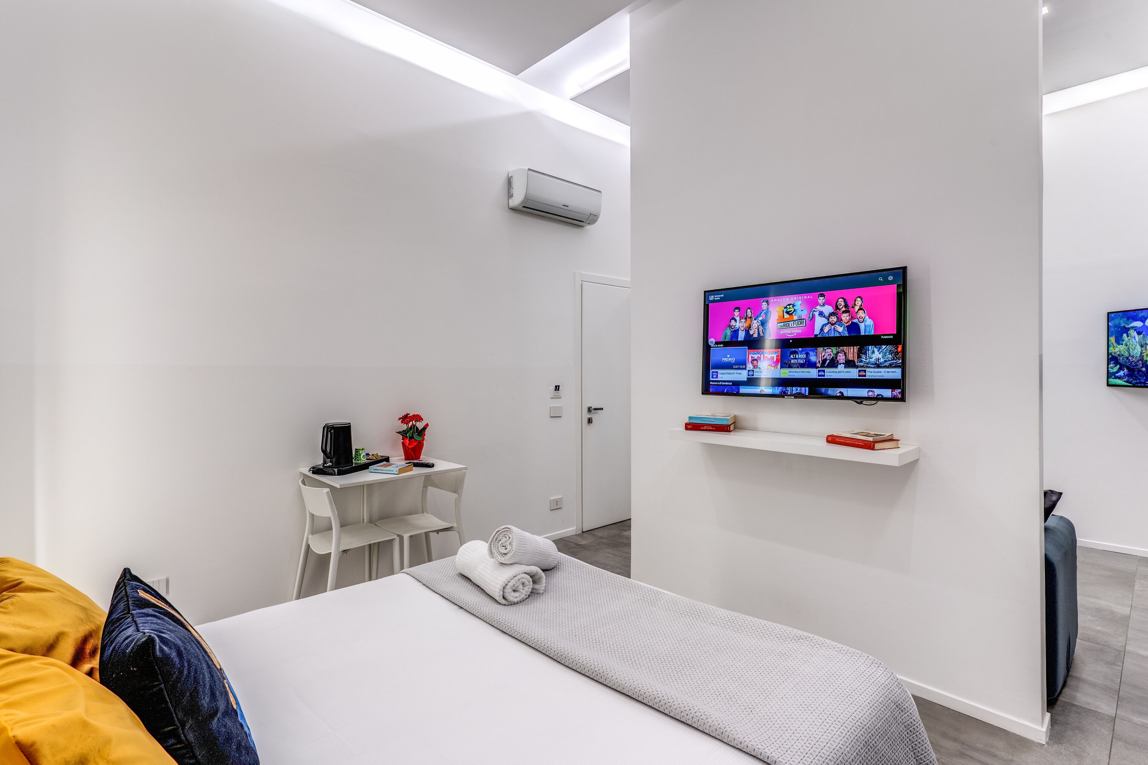 A modern hotel room with a bed, a TV mounted on the wall, a small desk with a chair, and a white wall.