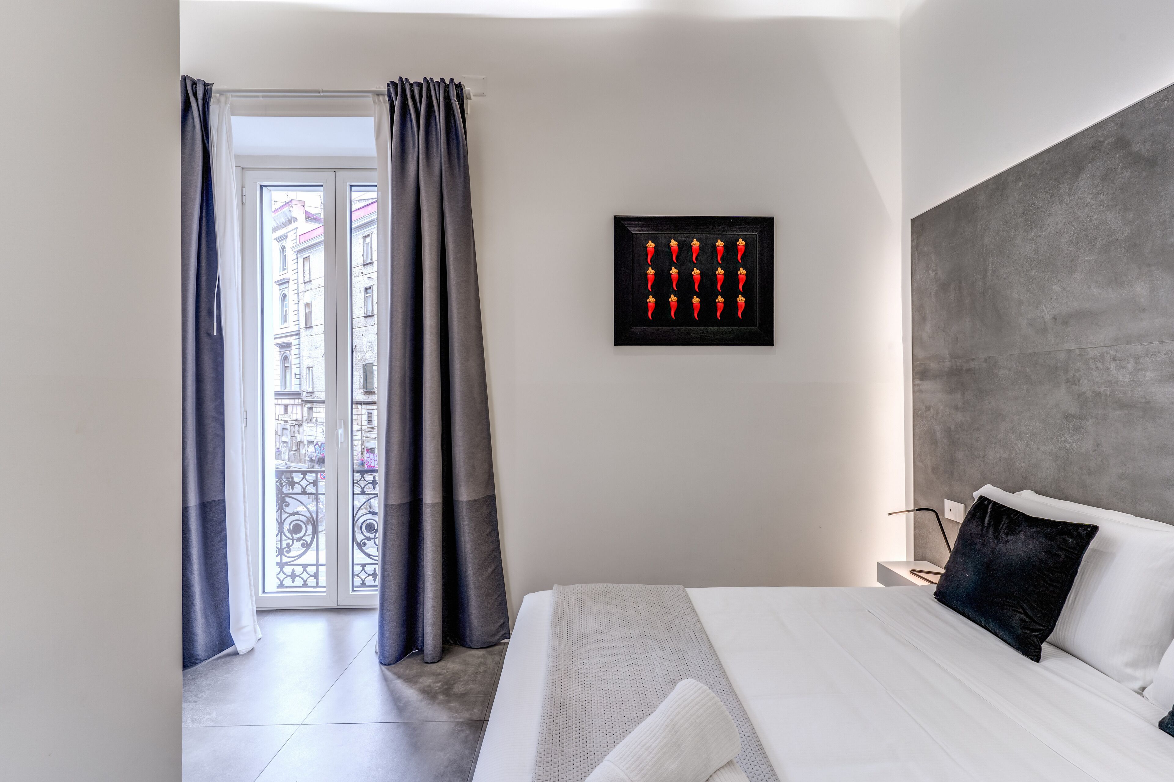 A modern hotel room with a large bed, a grey headboard, and a view of a cityscape through a balcony.