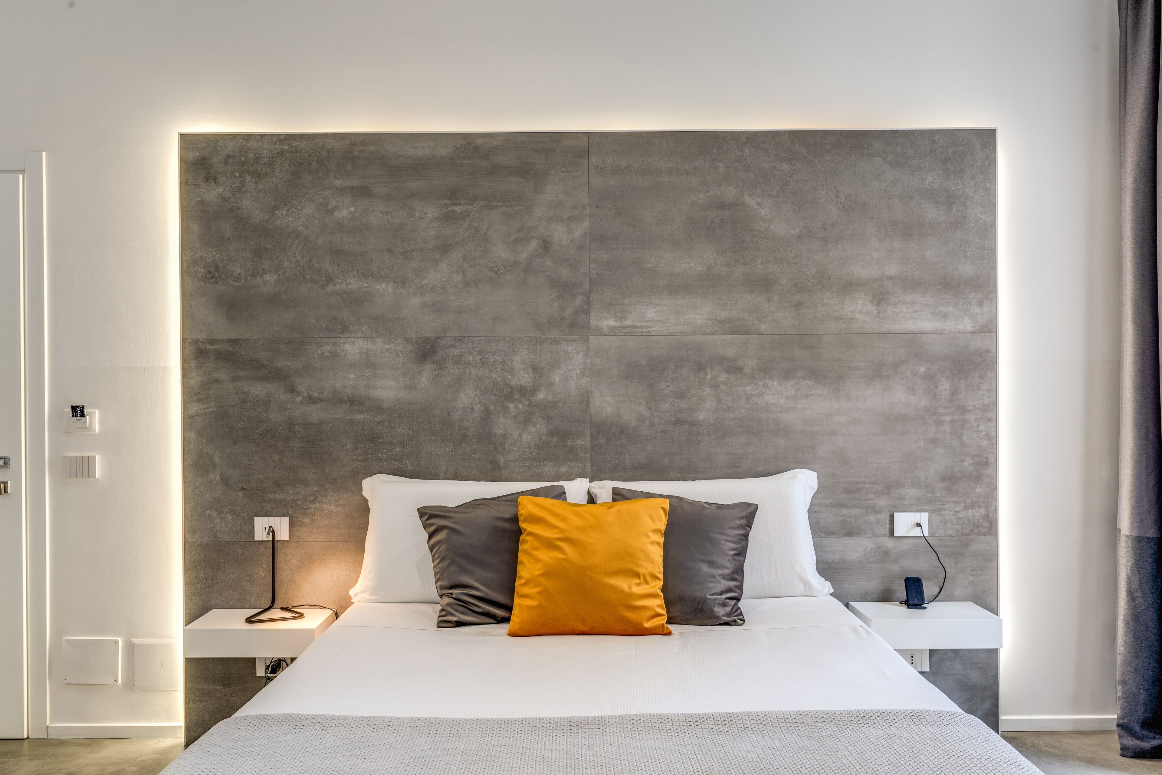 A modern hotel room with a large bed, a grey textured headboard, white bedding, and a bedside table with a lamp.