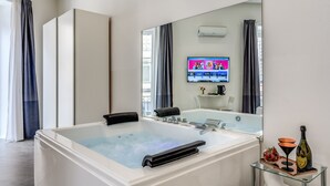 A modern bathroom with a large bathtub, a glass-topped vanity, and a flat-screen TV mounted on the wall. - Gallery Suite Naples (Naples)