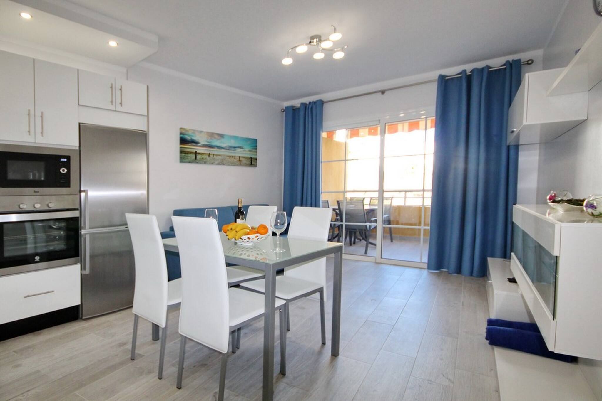A modern kitchen with white cabinets, a stainless steel refrigerator, and an oven. There is a dining table with white chairs and a bowl of fruit.