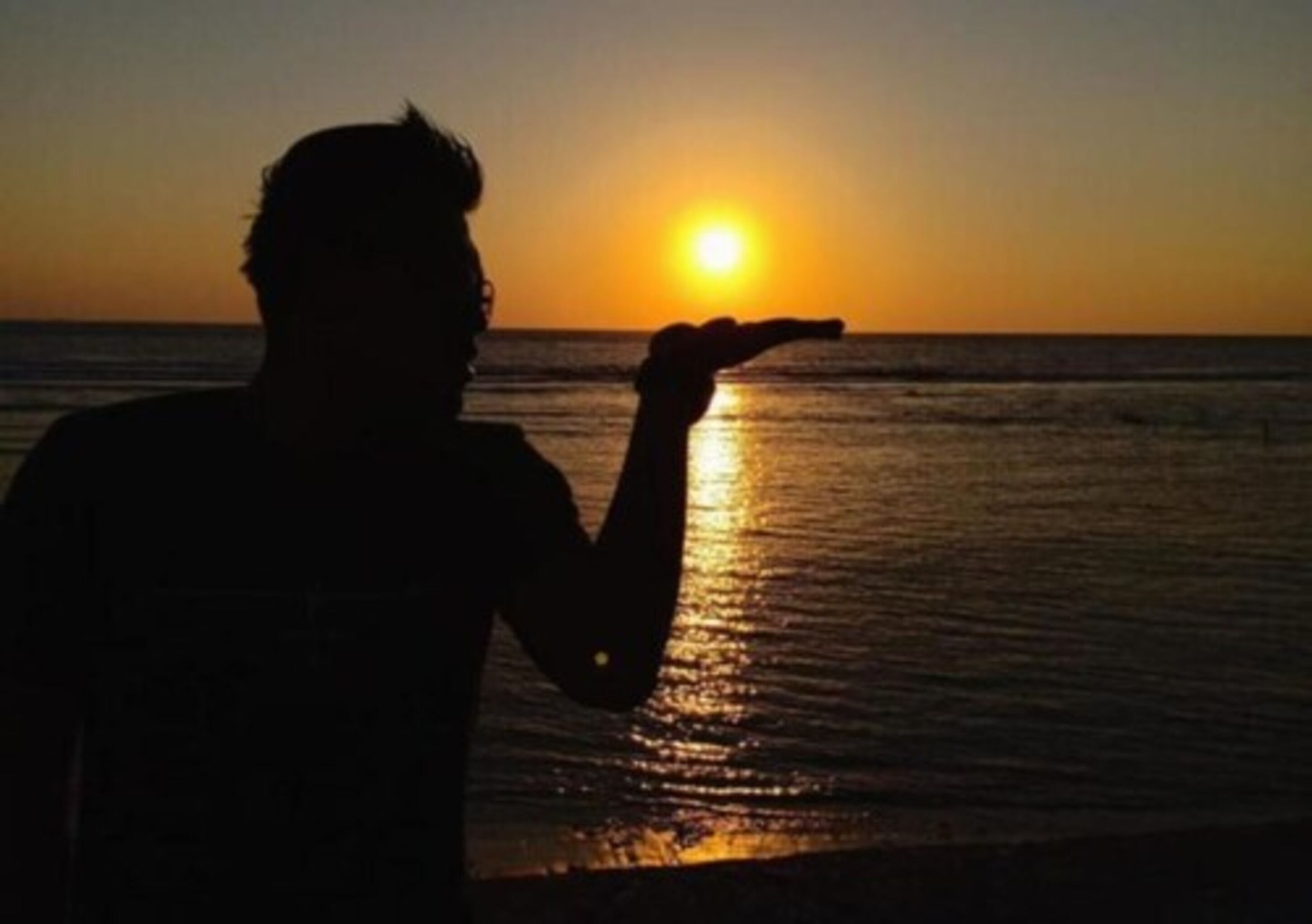A silhouette of a person holding their hand up against a sunset over the ocean.