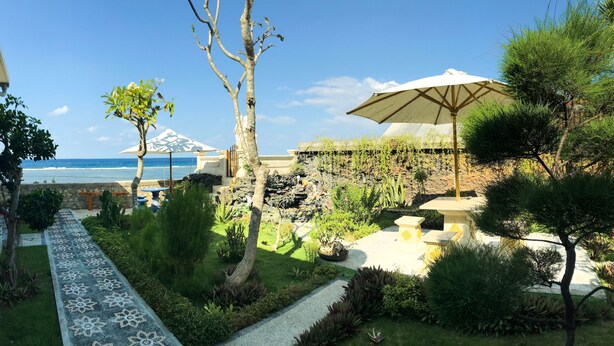 A garden with a pathway leading to outdoor seating, a view of the ocean, and a building with a white umbrella. - Swan Beach Inn (Penida Island)