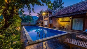 A swimming pool with wooden deck and lounge chairs, surrounded by lush greenery and a traditional building with a tiled roof. - The Amrta Borobudur (Borobudur)