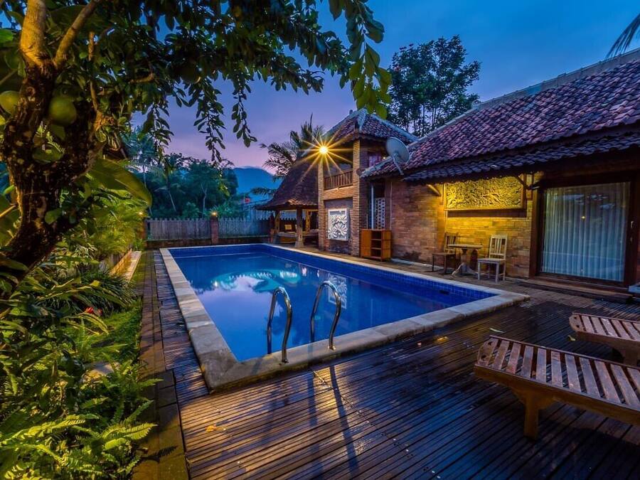 Piscina con muelle de madera y sillas de descanso, rodeada de exuberante vegetación y un edificio tradicional con tejado de baldosas.