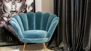 A teal upholstered chair with gold legs, positioned in front of a dark curtain and a large framed artwork. - Artale 07 - Adults Only (Split)
