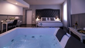 A modern hotel room with a large bathtub, a neatly made bed, a desk with a lamp, and a dining area with a bottle of champagne. - Villa Elisio Hotel & Spa (Naples)