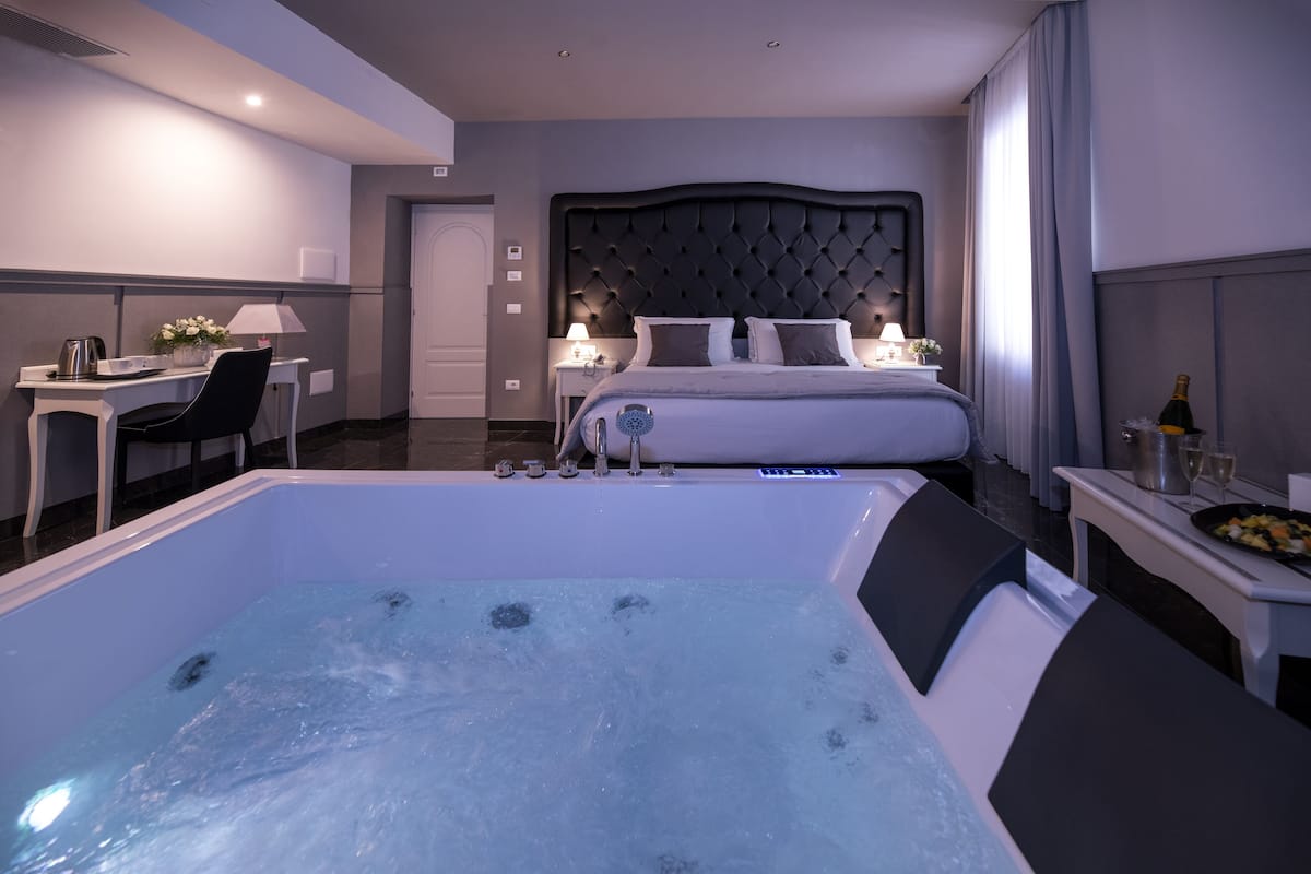 A modern hotel room with a large bathtub, a neatly made bed, a desk with a lamp, and a dining area with a bottle of champagne.