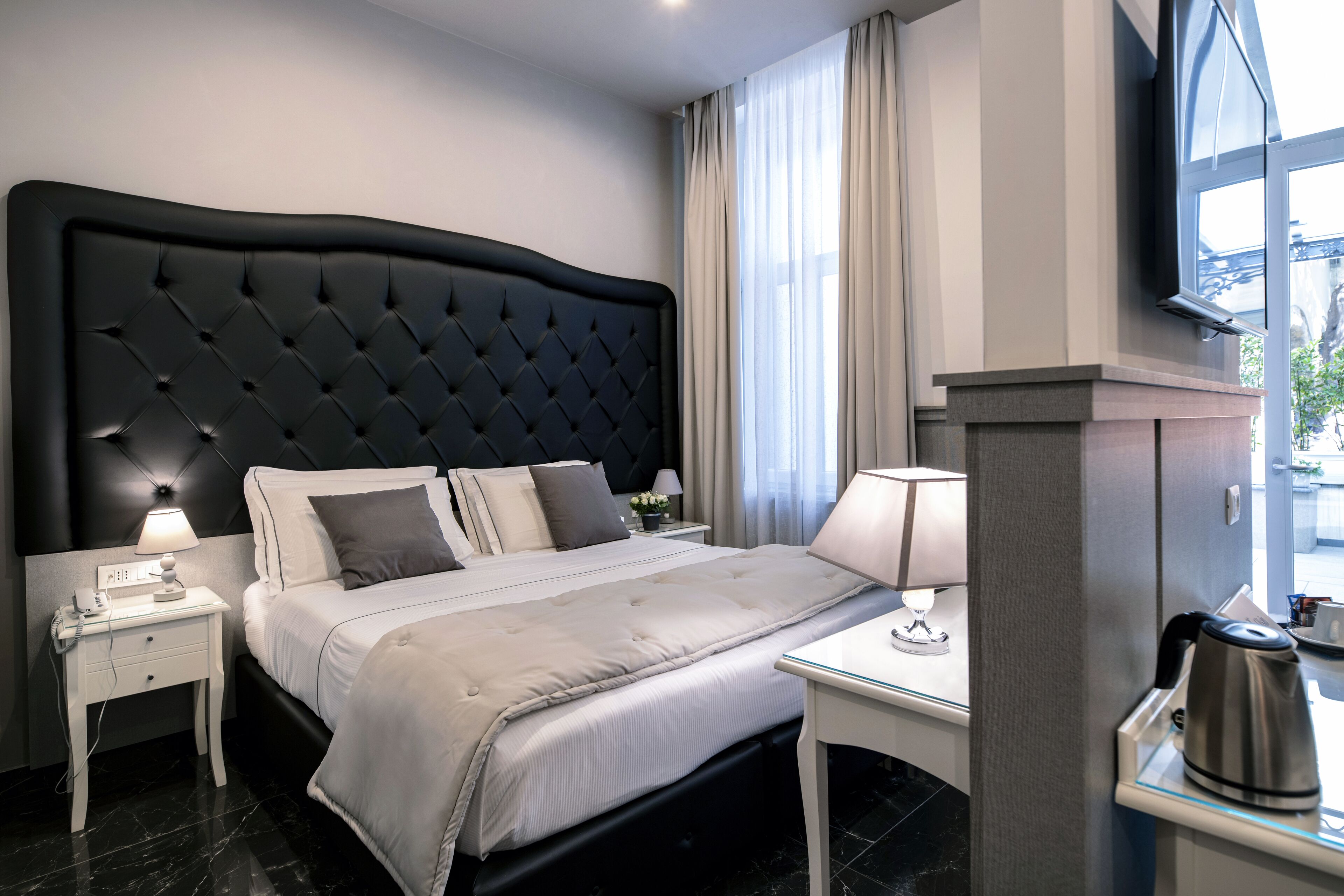 Ein modernes Hotelzimmer mit einem großen Bett, einem Nachttisch mit Lampe, einem kleinen Tisch mit Wasserkocher und einem Fenster mit Vorhängen.