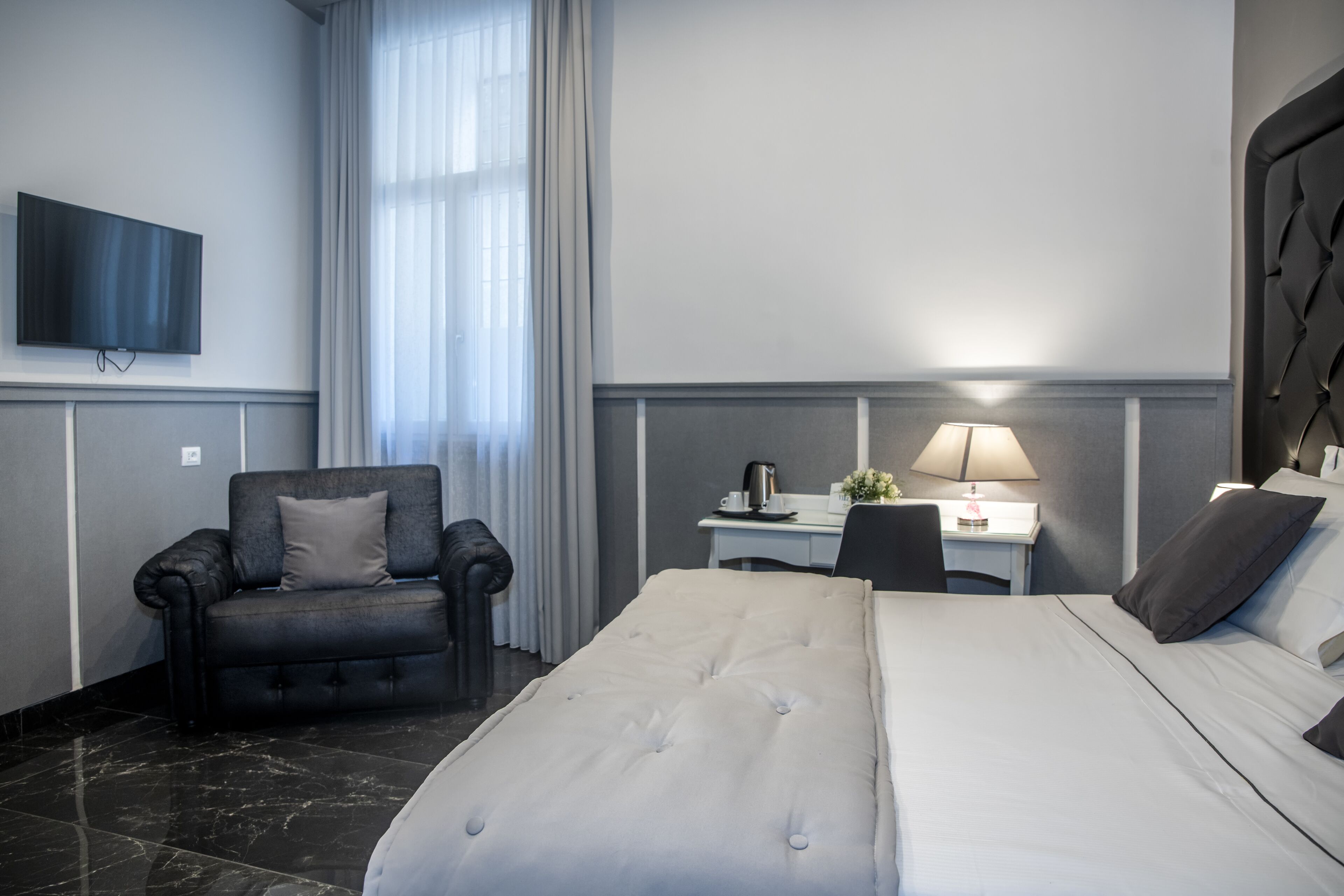 A hotel room with a large bed, a desk with a lamp, a chair, a TV mounted on the wall, and a window with curtains.