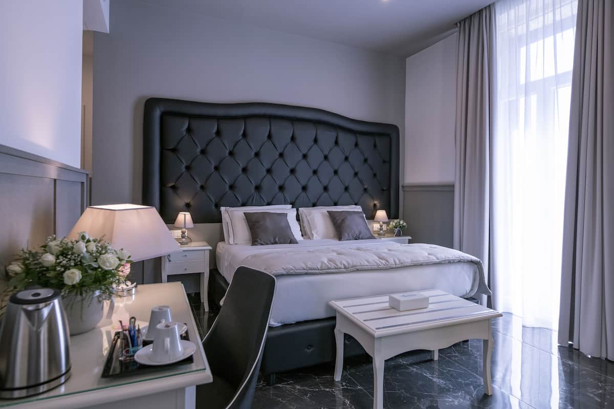 A hotel room with a large bed, a bedside table with a lamp, a small desk with a chair, and a coffee maker.
