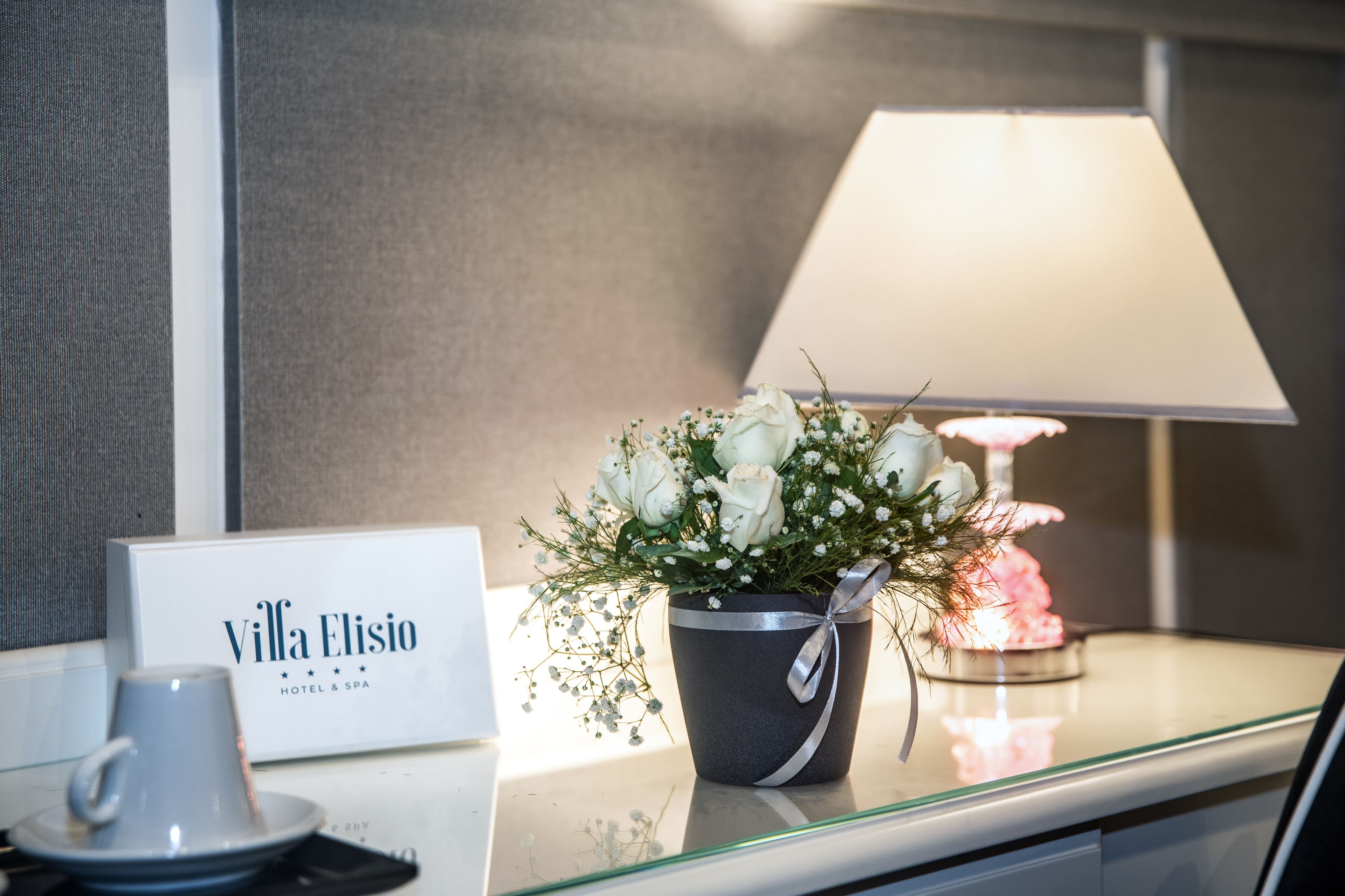 A hotel reception area with a sign reading "Villa Elisio HOTEL & SPA," a table with a floral arrangement, a lamp, and a cup and saucer.