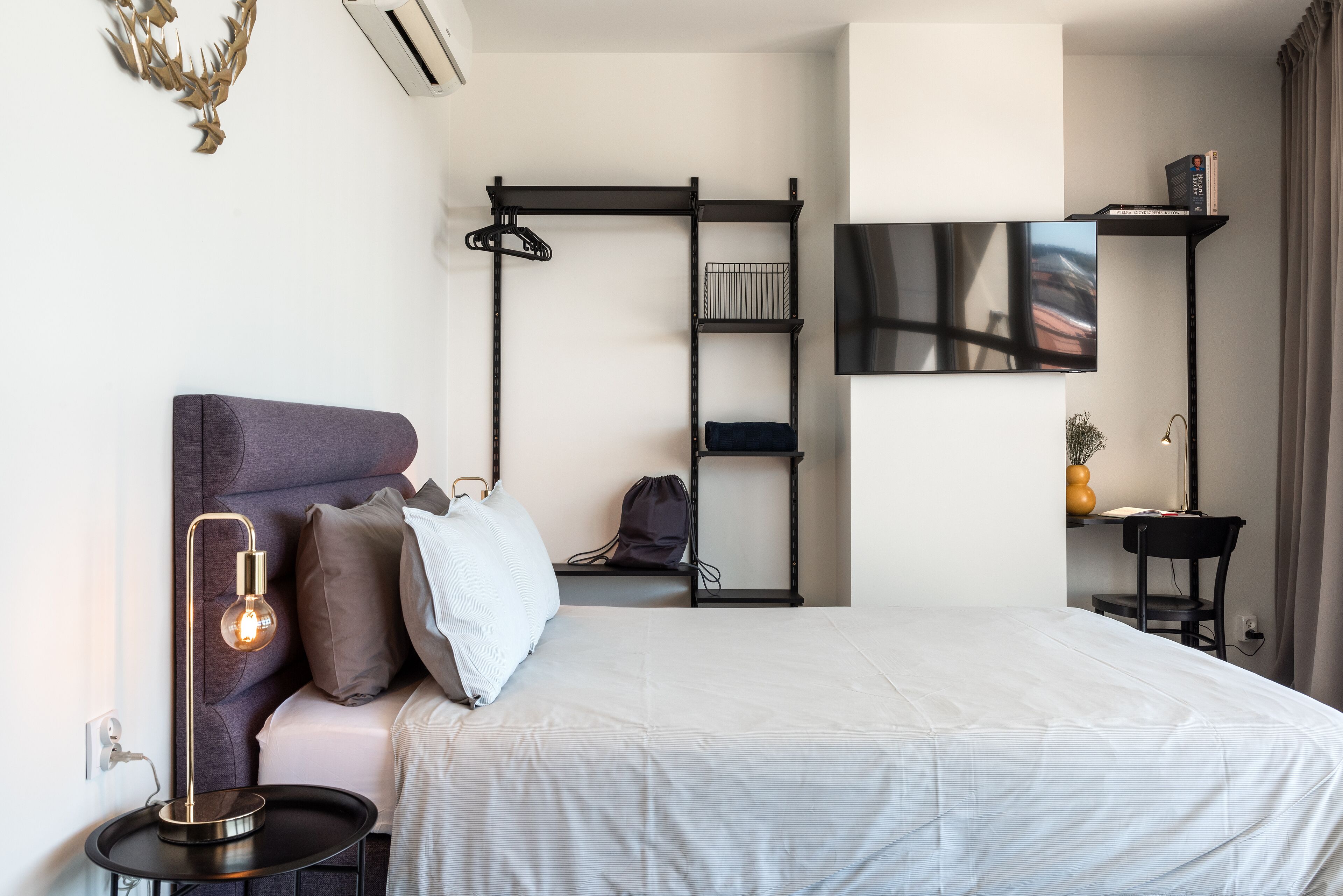 Quarto de hotel moderno com cama, mesa de cabeceira, luminária, TV e prateleira na parede.