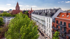 A row of modern buildings with balconies, a church with a tall spire, and a tree in the foreground.