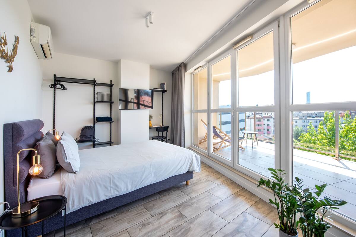 Ein modernes Schlafzimmer mit einem großen Bett, einem Nachttisch mit Lampe, einer Wandregal und Blick auf die Stadt durch große Fenster.
