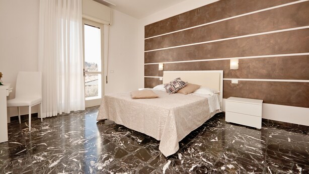 A modern bedroom with a bed, nightstands, a chair, and a large window with curtains. - Appartamento Anna (San Felice del Benaco)