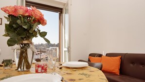 A dining table set with plates, cutlery, and a vase of flowers, with a view of a cityscape through an open window. - Appartamento Anna (San Felice del Benaco)