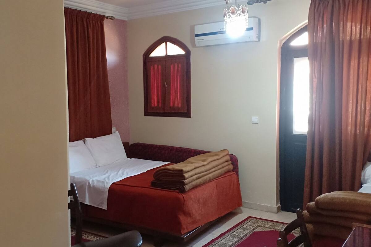 Un dormitorio con cama, ventana con cortinas, equipo de aire acondicionado y una puerta.