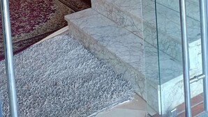Una escalera con escalones de mármol y barandilla de cristal, que conduce a una habitación con alfombra estampada y sillas de madera.