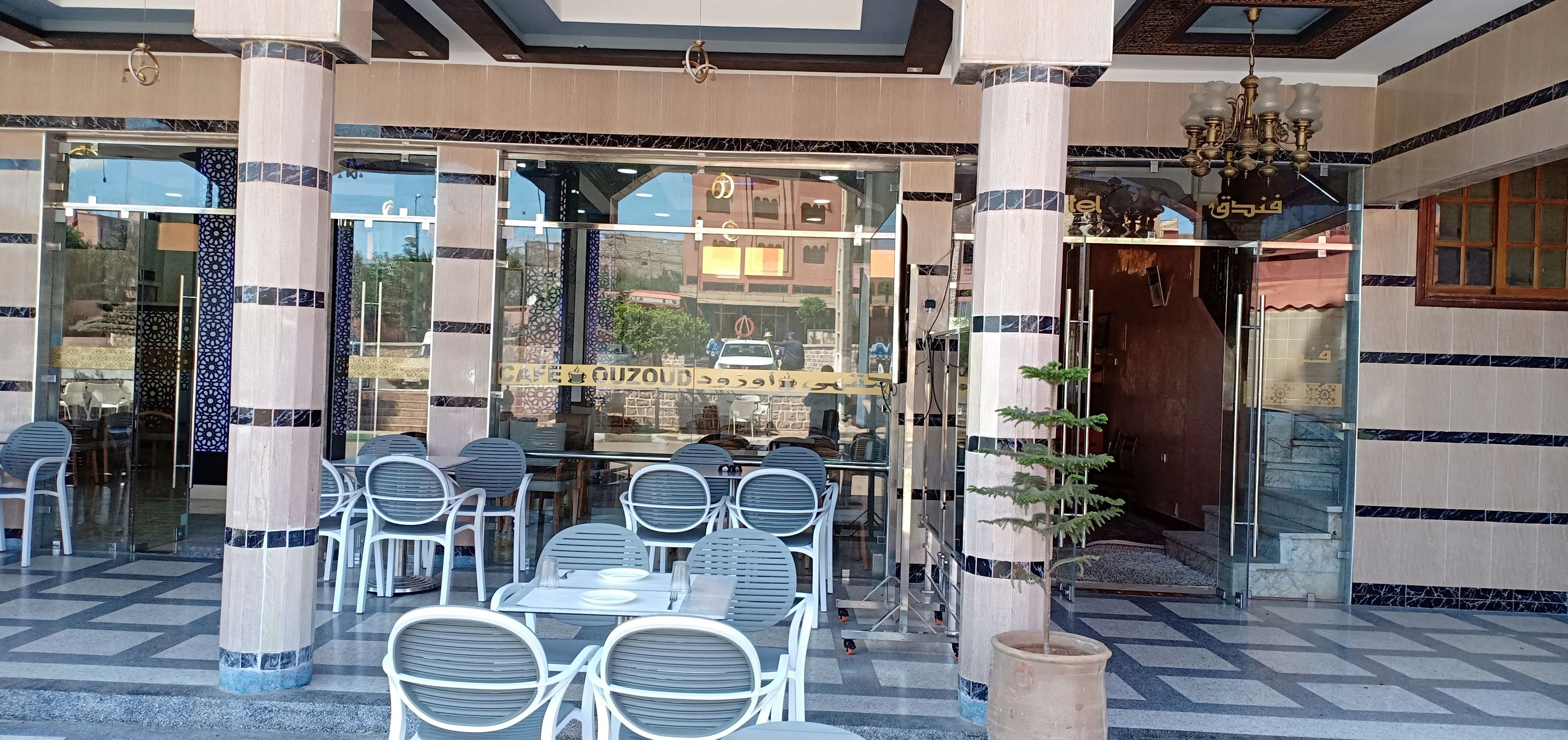 A hotel entrance with a glass facade, outdoor seating area, and a sign reading "CAFE GUZOUD".