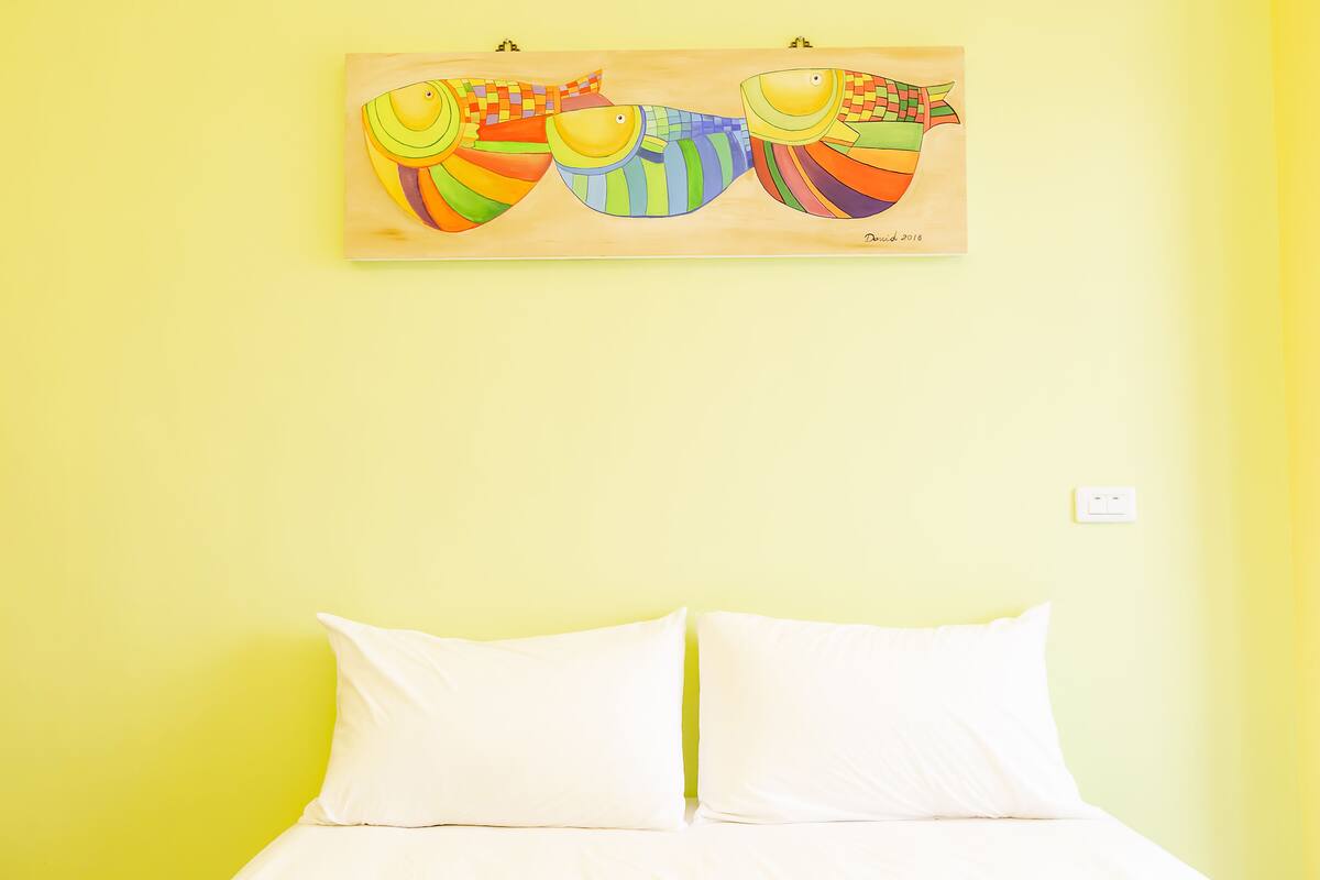 Una cama con ropa blanca sobre un pared amarillo, una obra de arte colorida encima y un interruptor de luz en la pared.