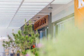 Entrada de hotel con letrero de madera, puertas de cristal y un área exterior cubierta con plantas y una mesa.