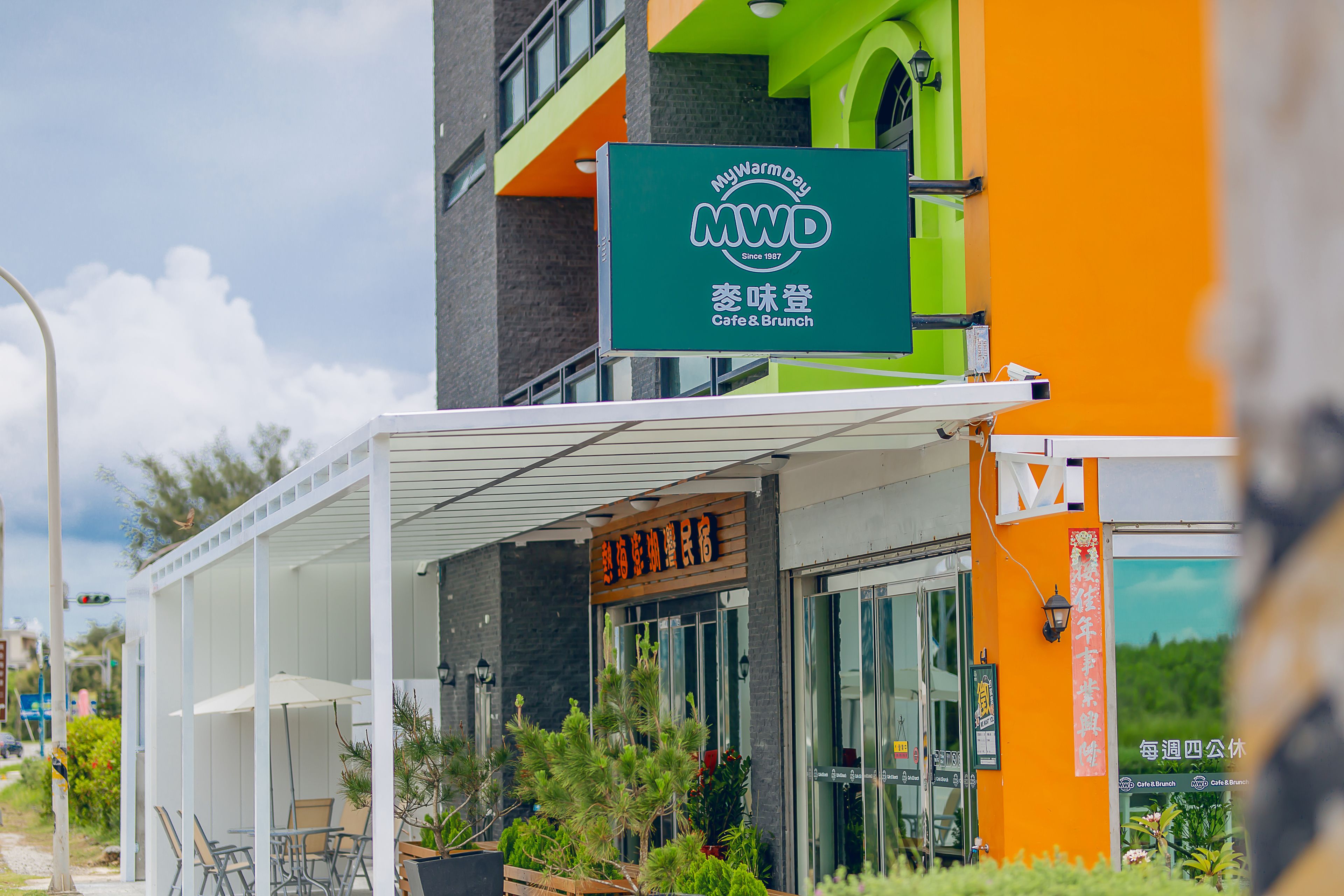 A cafe with outdoor seating, a green sign, and a modern building facade.
