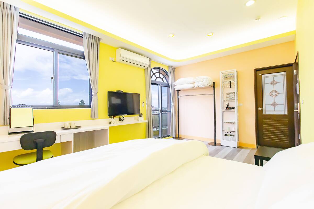 Habitación de hotel con cama, escritorio con silla, televisor, ventana con cortinas y puerta.