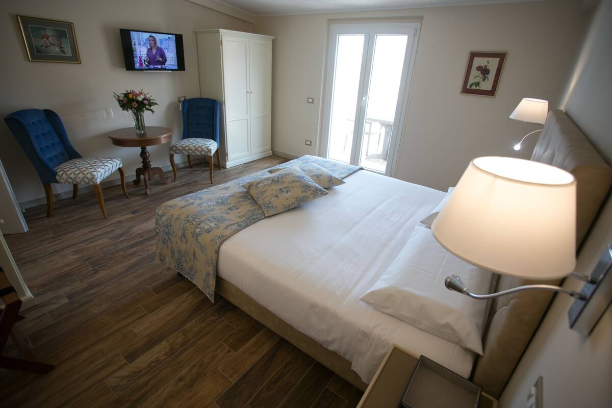 A bedroom with a bed, a TV, a chair, a table with flowers, and a lamp.