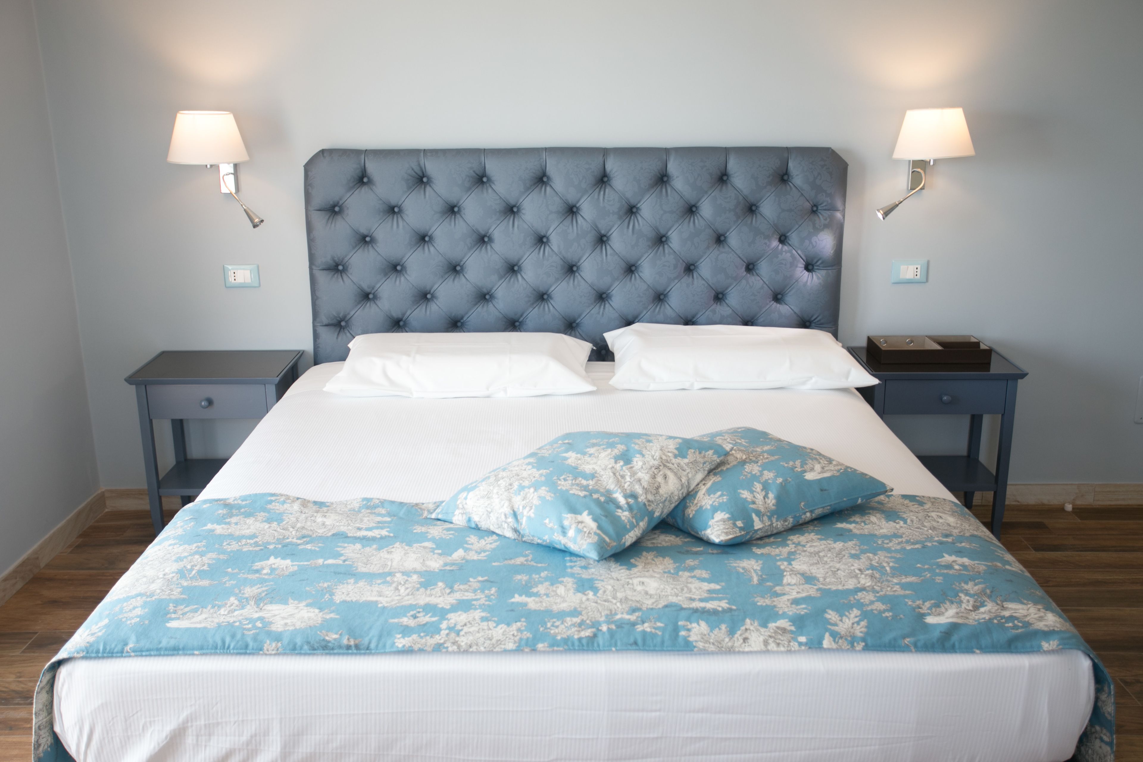 Un dormitorio con cabecera capitonada, cama con ropa de cama floral en blanco y azul, dos mesitas de noche con lámparas y una mesita con lámpara.