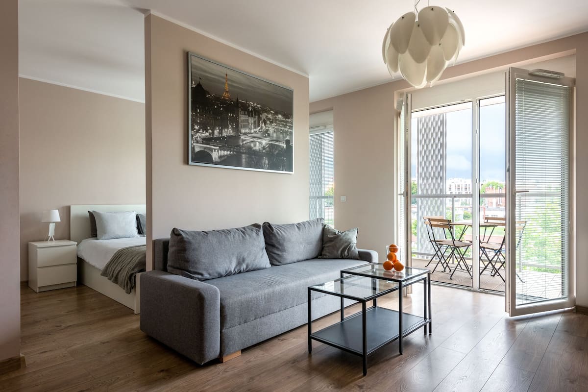 A modern living room with a grey sofa, a glass coffee table, a bed with a white headboard, and a cityscape painting on the wall.