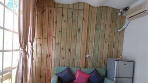 A room with a wooden paneled wall, a green sofa with colorful cushions, a small refrigerator, and a window with curtains. - Blue Pa Hotel (Hengchun)