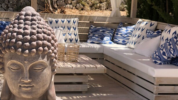 Eine Buddha-Statue in einem Garten mit einer Steinmauer, hölzernen Bänken und gemusterten Kissen.