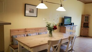Un coin repas avec une table en bois, des chaises, un banc avec des coussins, un vase avec des fleurs et un téléviseur posé sur un meuble.