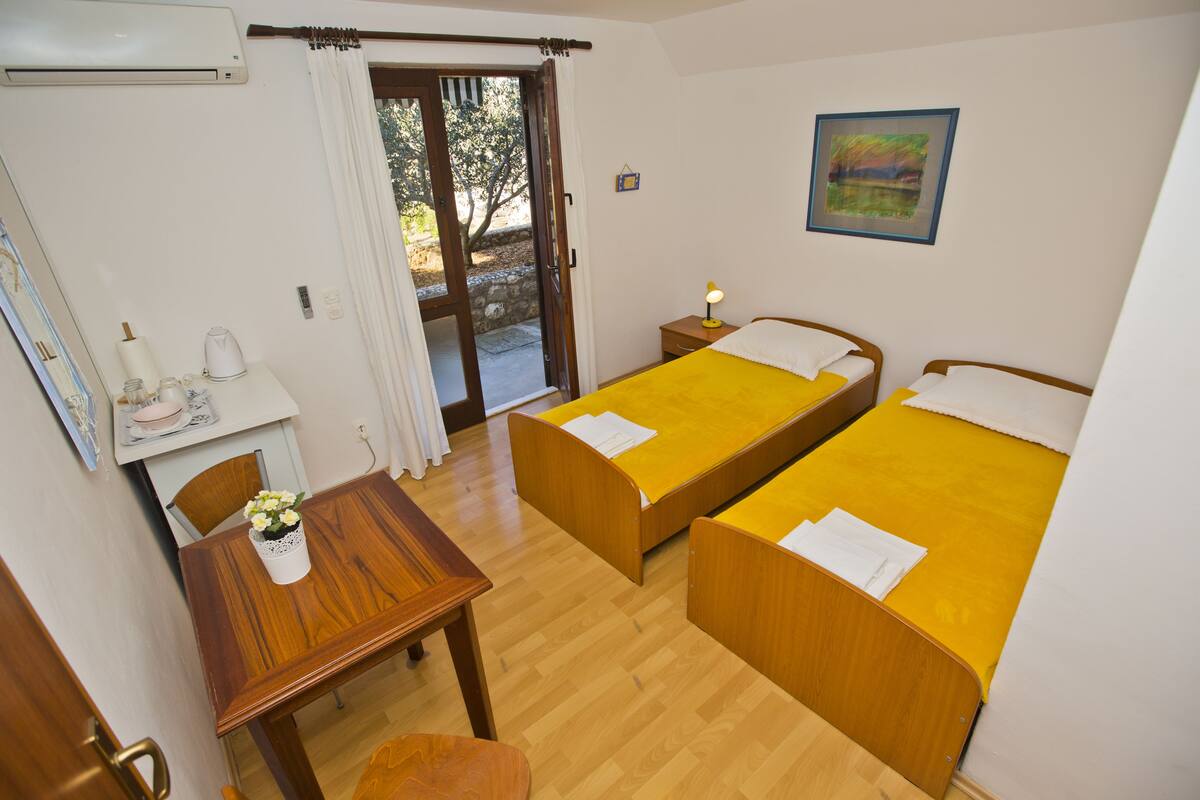 A hotel room with two single beds, a wooden table, a small desk with a chair, a window with curtains, and a painting on the wall.