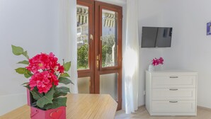 A room with a wooden table, a white dresser, a TV mounted on the wall, and a window with a view of buildings and trees. - Guest House Kovacevic (Hvar)