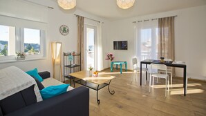 A modern living room with a sofa, coffee table, and dining table. There are curtains, a TV, and a view of the outdoors. - Guest House Kovacevic (Hvar)