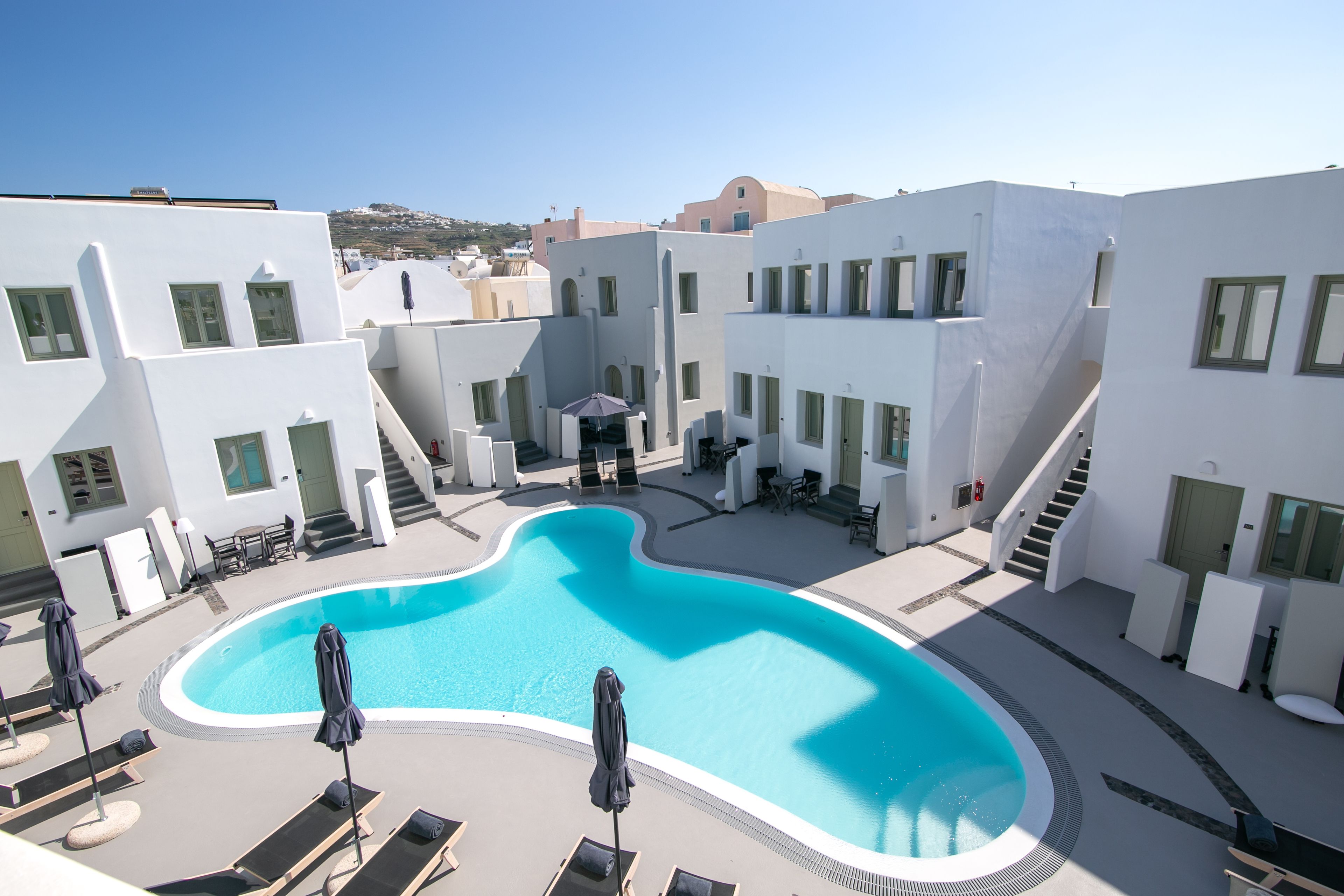 A pool area with lounge chairs and umbrellas, surrounded by white buildings with multiple windows and a staircase.
