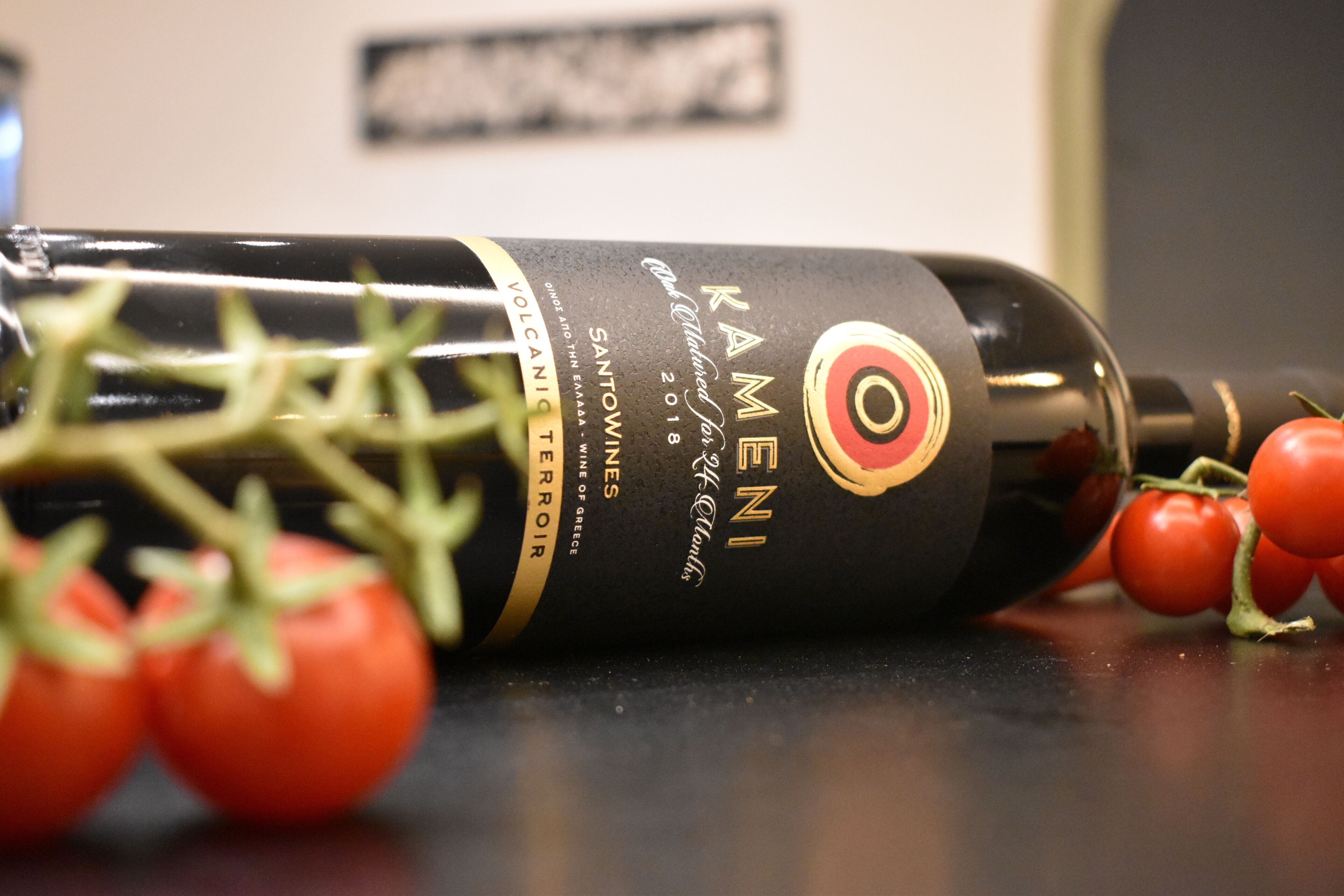 Una botella de vino Kamenis Santorini con etiqueta de diseño rojo y dorado, sobre una superficie con algunos tomates al frente.