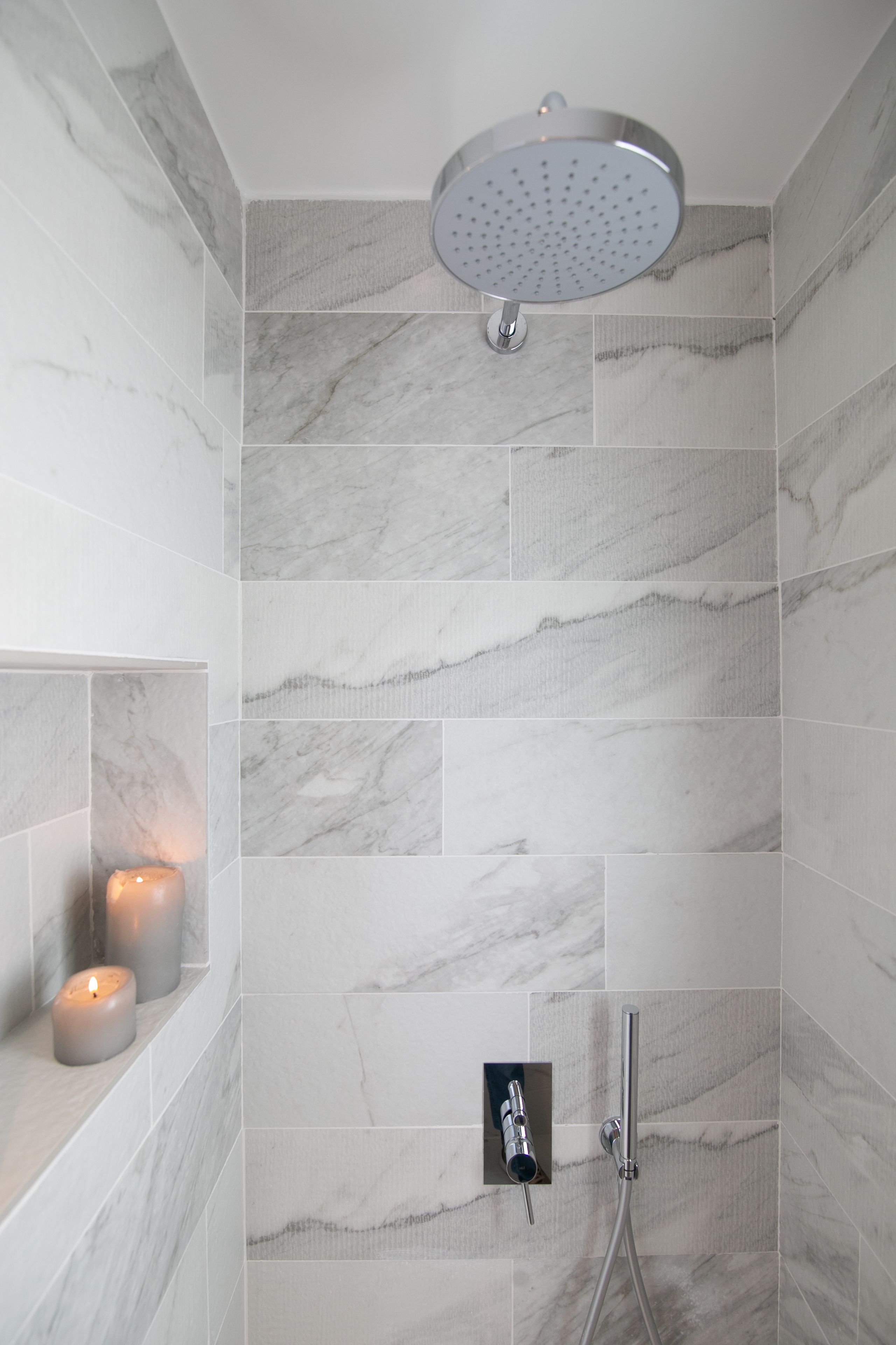 Una ducha moderna con cabezal de ducha de mano, grifo empotrado en la pared y una zona de ducha revestida con azulejos que imitan el mármol.