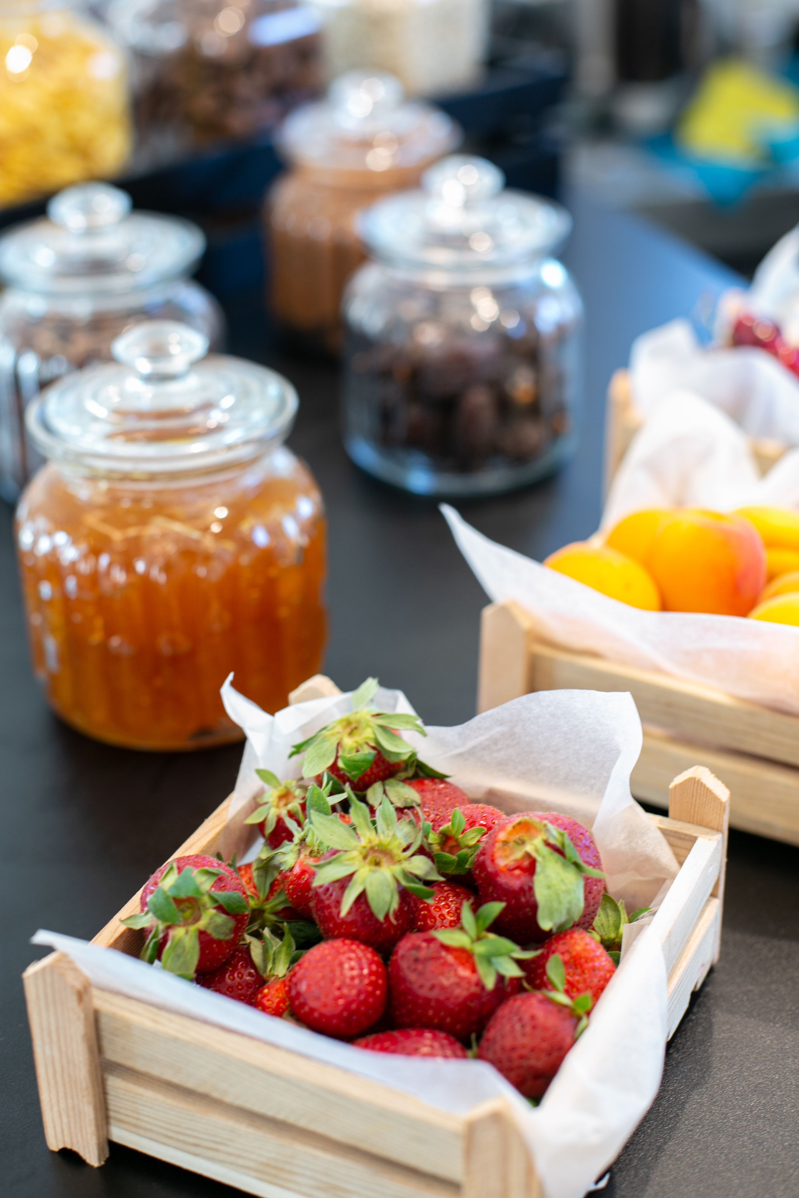 Una caja de madera llena de fresas frescas, un tarro de miel y una cesta de naranjas sobre una mesa.