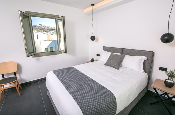 Una habitación de hotel moderna con una cama grande, vistas a través de una ventana abierta, una silla y una mesita de noche con una planta.
