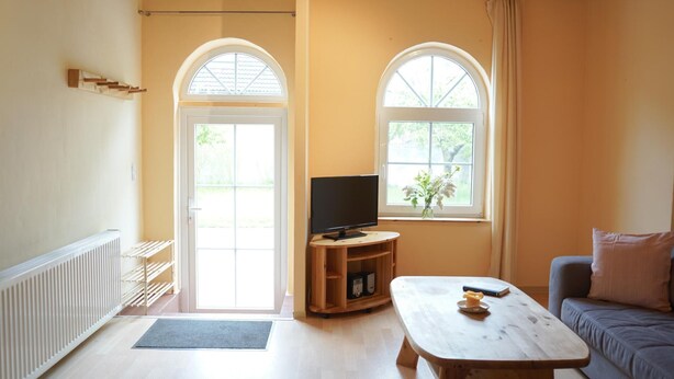 A living room with a television on a wooden stand, a sofa, a coffee table, and two windows with curtains. - Ferienwohnung Muschel (Kopendorf auf Fehmarn)
