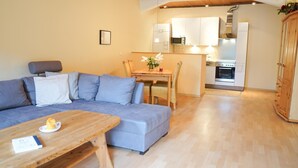 A modern living room with a grey sofa, a wooden coffee table, and a dining area with a wooden table and chairs. A kitchen with white cabinets and stainless steel appliances is visible in the background. - Ferienwohnung Muschel (Kopendorf auf Fehmarn)